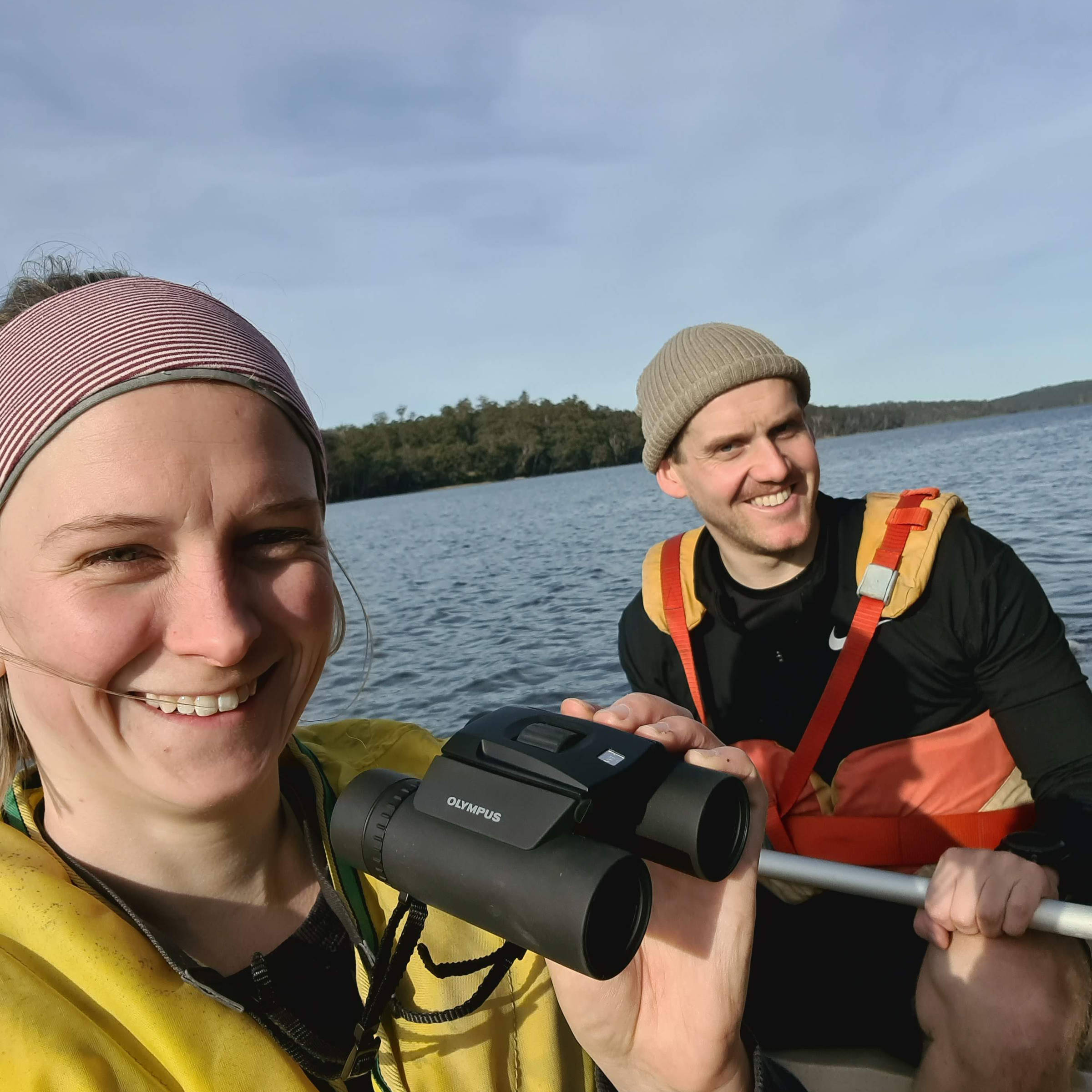 A man and woman in their twenties, wearing life jackets and sitting in a kayak on water, with land in the distance, smile at the camera. The woman is holding up a pair of binoculars. Photo: Kelsey Picard.