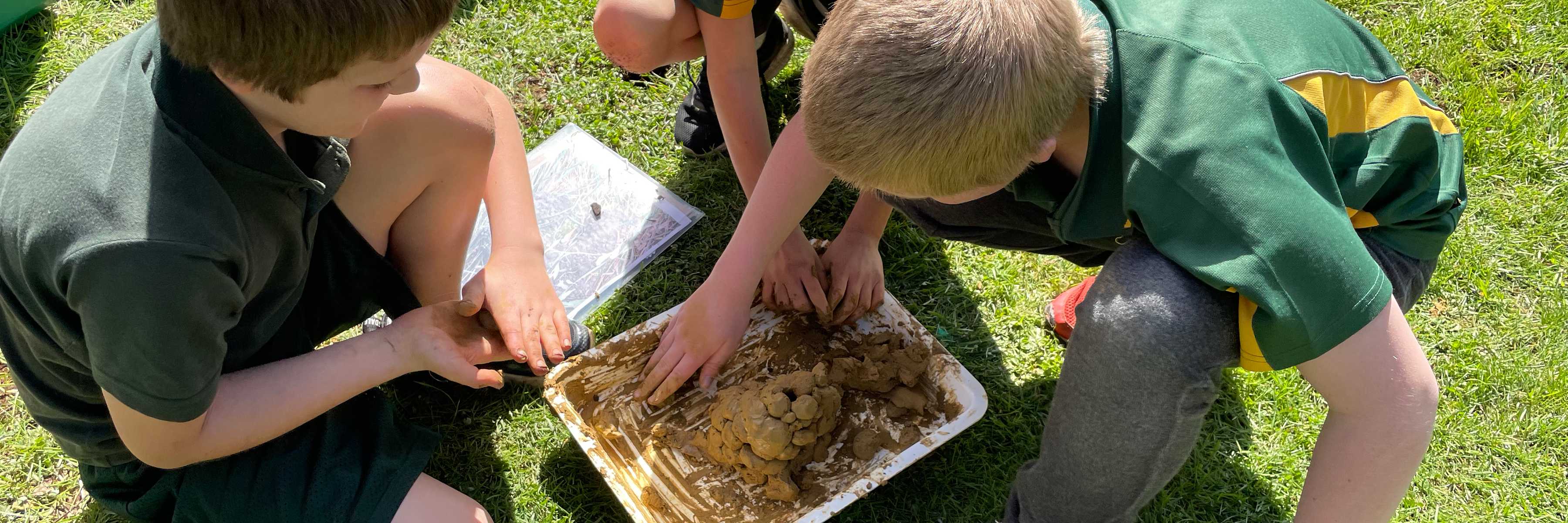 Three students from Sassafras Primary School work with clay in a white tray sitting on short green grass. Photo: Clare Hawkins.