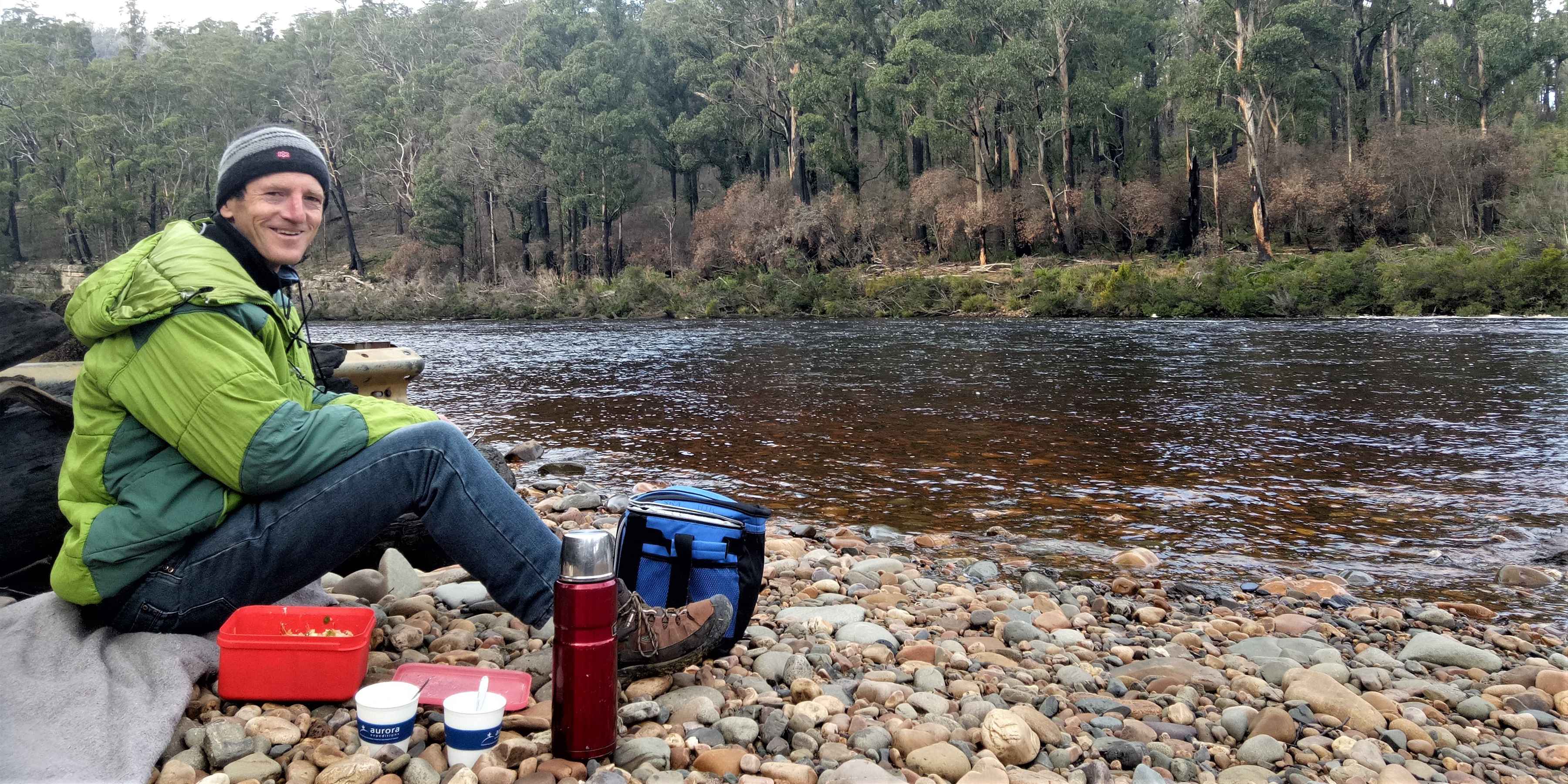A warmly-dressed man sitting on stream-rounded pebbles at the edge of a broad river smiles at us. He has a thermos, two cups and other picnic material beside him.
