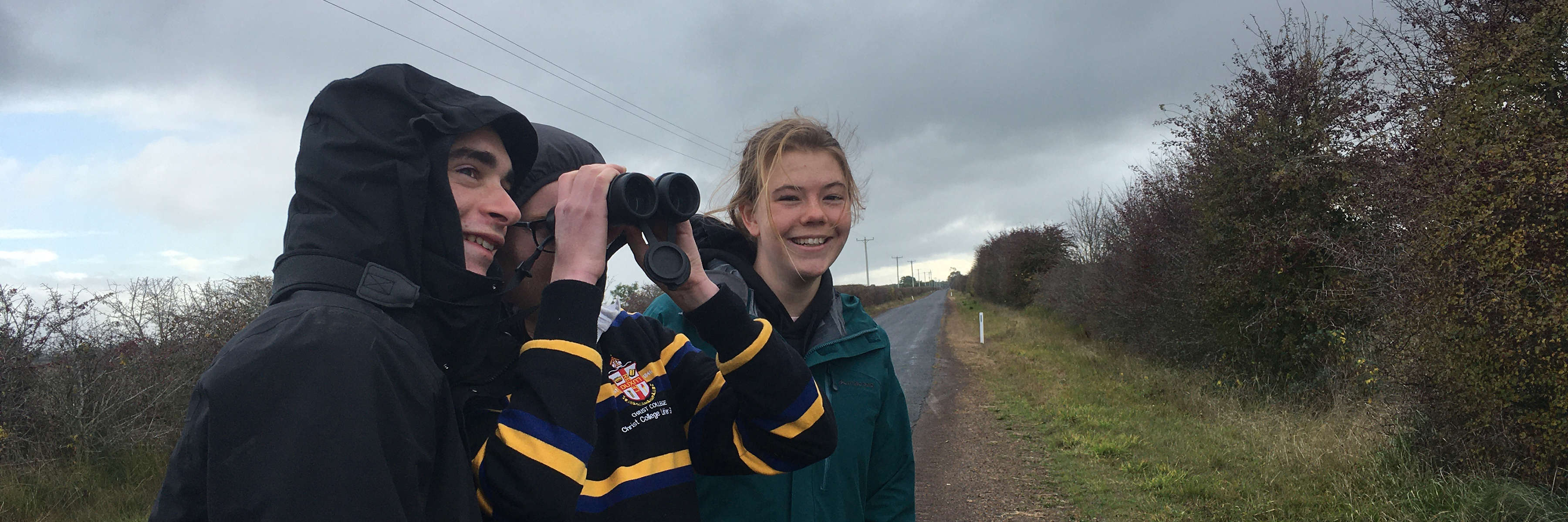 Three university students on a roadside in an agricultural area pose for the camera after a 10-minute Where? Where? Wedgie! survey. One of them is looking to the sky with binoculars. Photo: Katherine Stuart.