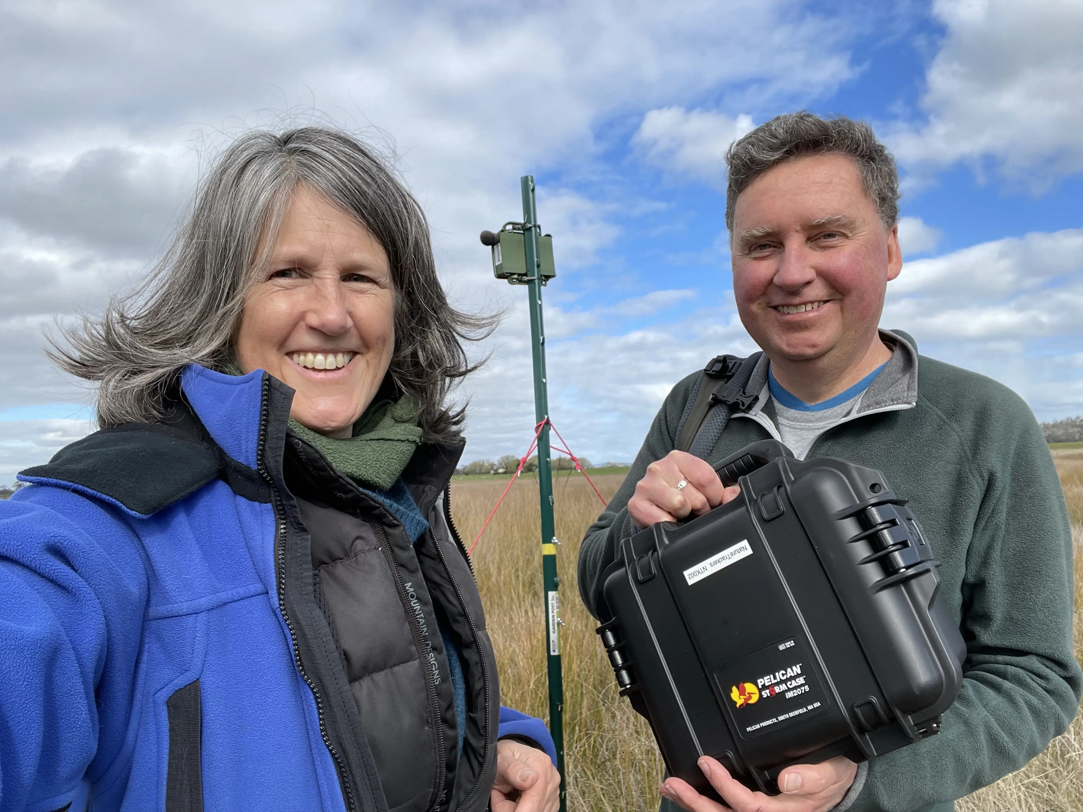 Clare Hawkins and Jim Lovell selfie with a small green acoustic recorder set up high on a stake behind them, at a wetland. Photo: Clare Hawkins.