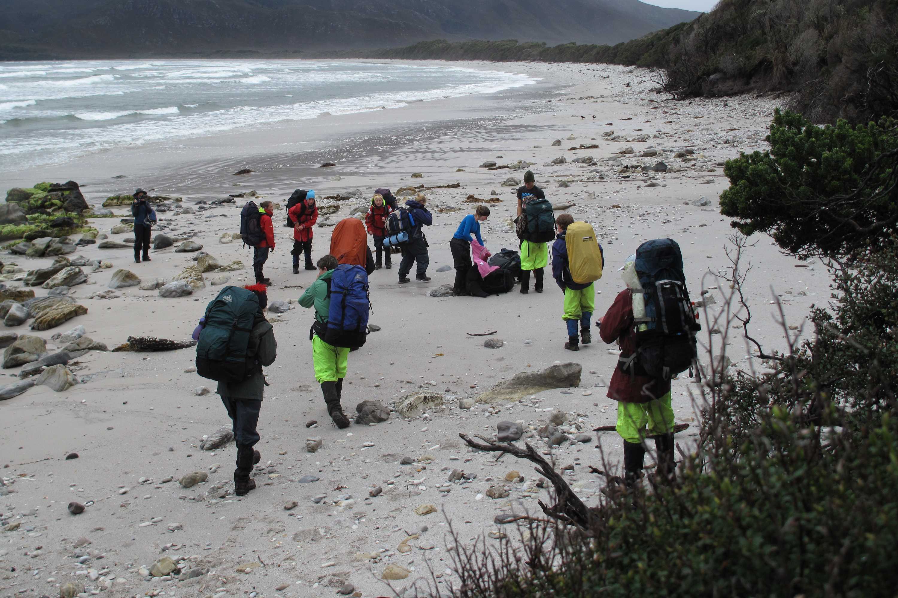A group of people with large backpacks walk along a white sandy beach on a dull day, with hills rising up in the background. Photo: Andrew Hughes.
